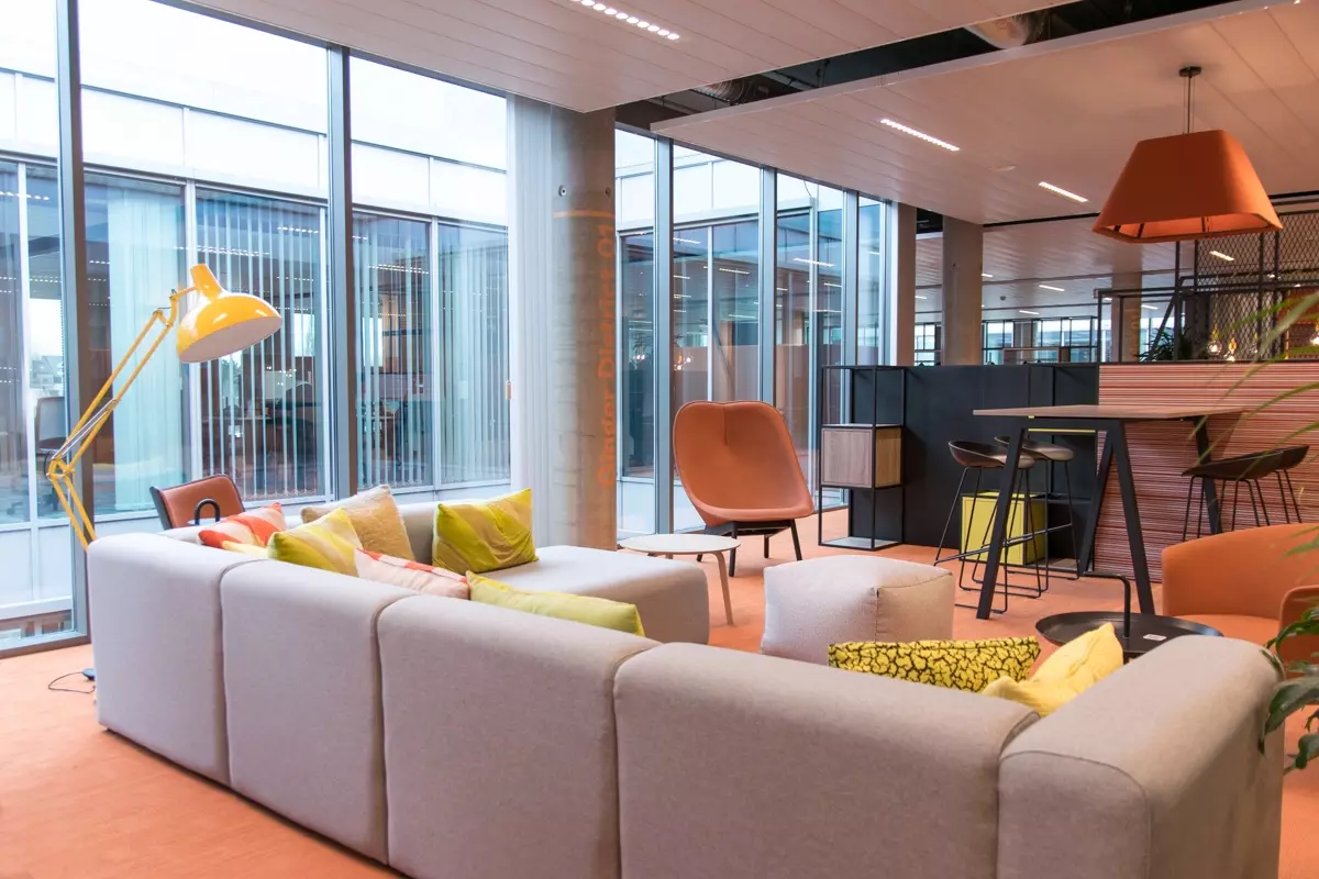 Een open kantooromgeving met een comfortabele zithoek, met een grijze bank, kleurrijke kussens, en oranje zetels.