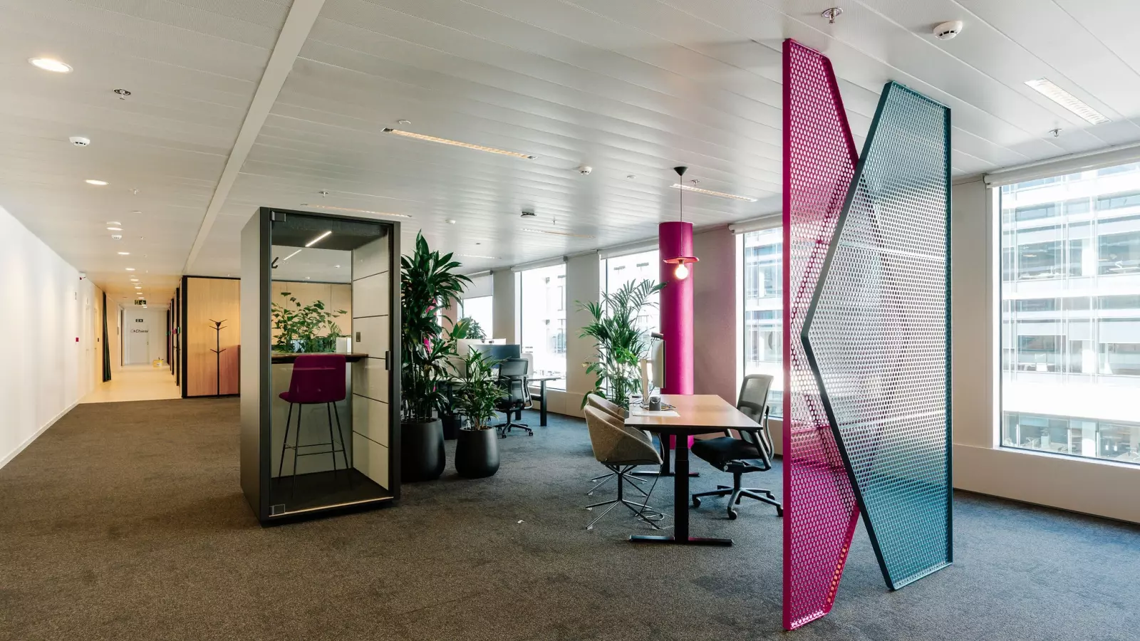 Modern office with a colourful room divider, a black phone booth and plants near the workstations and desks.